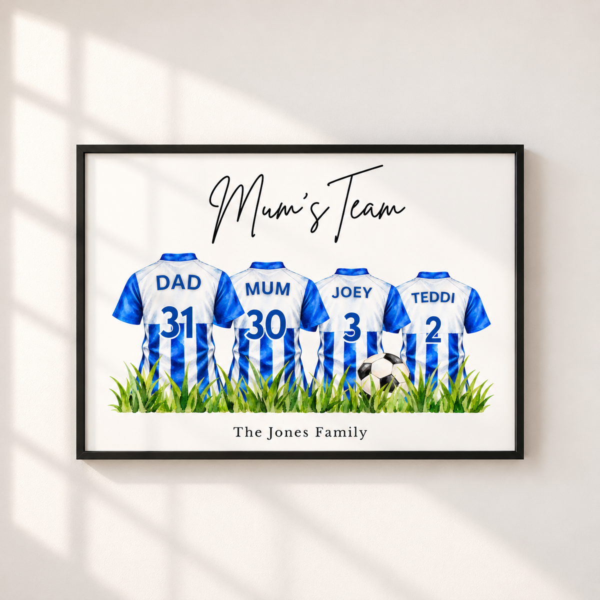 A framed personalized wall art print showing four soccer jerseys labeled DAD, MUM, JOEY, and TEDDI with numbers 31, 30, 3, and 2, above green grass and a small soccer ball. The print reads 'Mum's Team' and 'The Jones Family,' displayed on a light wall with soft window shadows.
