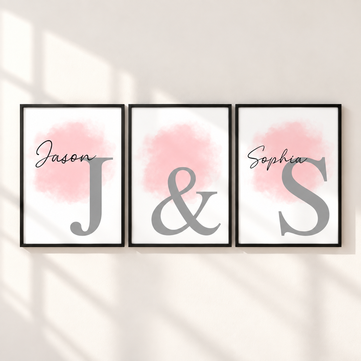 Three framed personalized wall art prints hang on a light wall with soft window-shadow lighting. Each print has a black frame, a white background, a soft pink watercolor wash, and a large gray letter: Jason, J, and S, with an ampersand between the middle and right prints and the name Sophia above the S.
