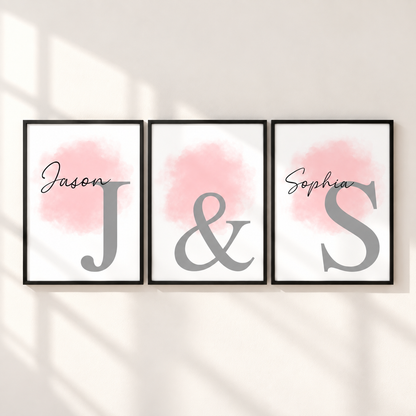 Three framed personalized wall art prints hang on a light wall with soft window-shadow lighting. Each print has a black frame, a white background, a soft pink watercolor wash, and a large gray letter: Jason, J, and S, with an ampersand between the middle and right prints and the name Sophia above the S.