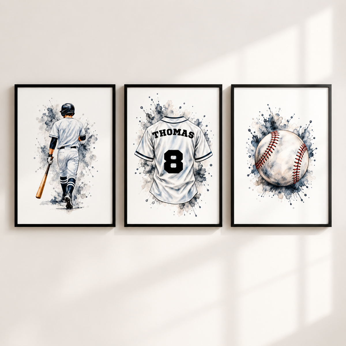 Three framed baseball-themed wall art prints hang on a light beige wall with soft window-shadow lighting. The left print shows a watercolor-style baseball player holding a bat, the center shows a white baseball jersey with the name THOMAS and number 8, and the right shows a baseball with red stitching over a dark blue watercolor splash.