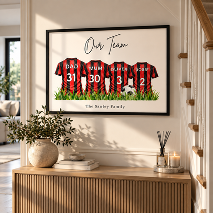 A framed personalized family jersey print hangs on a cream wall above a wooden console table. The artwork shows four red-and-black striped soccer jerseys with the names Dad, Mum, Oliver, and Rosie, plus numbers 31, 30, 3, and 2, under the text 'Our Team' and 'The Sawley Family'.