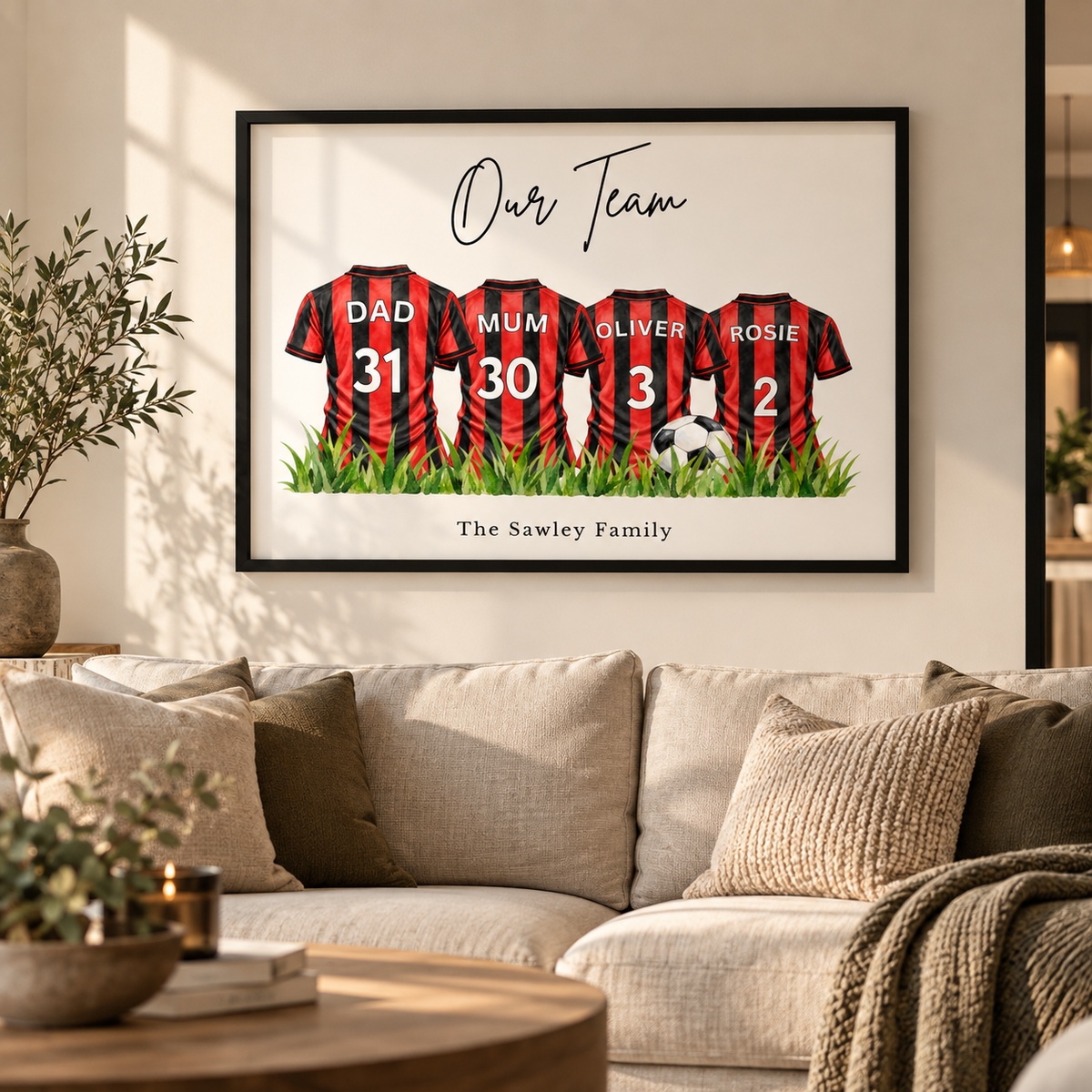 A framed personalized family soccer jersey wall art print hangs above a beige sofa in a styled living room. The print shows four red-and-black striped jerseys labeled Dad, Mum, Oliver, and Rosie with numbers 31, 30, 3, and 2, under the text 'Our Team' and above 'The Sawley Family'.