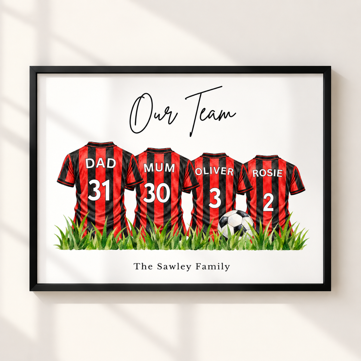 A framed personalized wall art print showing four red-and-black striped soccer jerseys with the names Dad, Mum, Oliver, and Rosie, plus the text 'Our Team' and 'The Sawley Family'. The framed print is displayed against a light wall with soft window shadows.