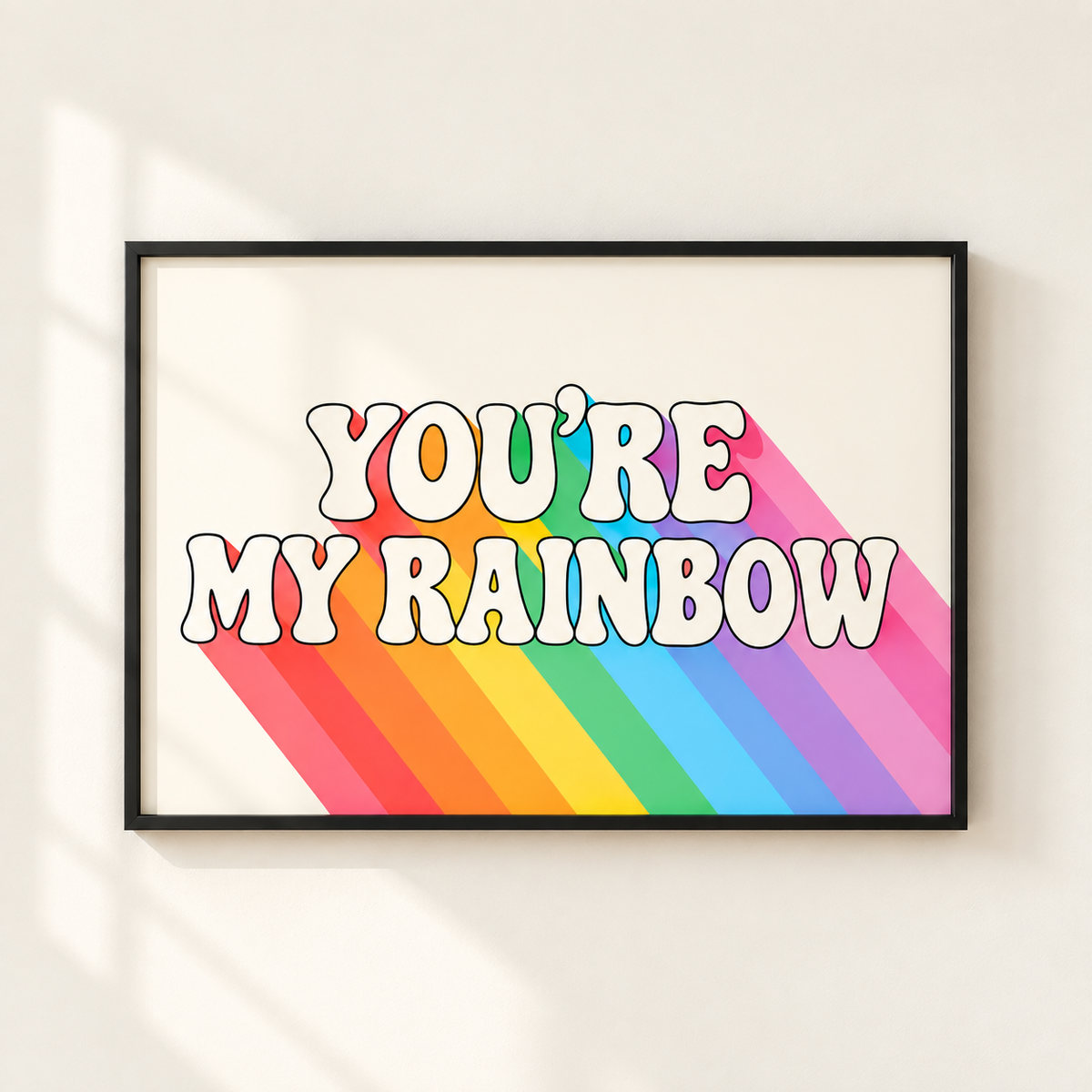 A framed wall art print with the phrase “YOU’RE MY RAINBOW” in large white bubble letters over diagonal rainbow stripes, shown hanging on a light beige wall with soft window shadows.