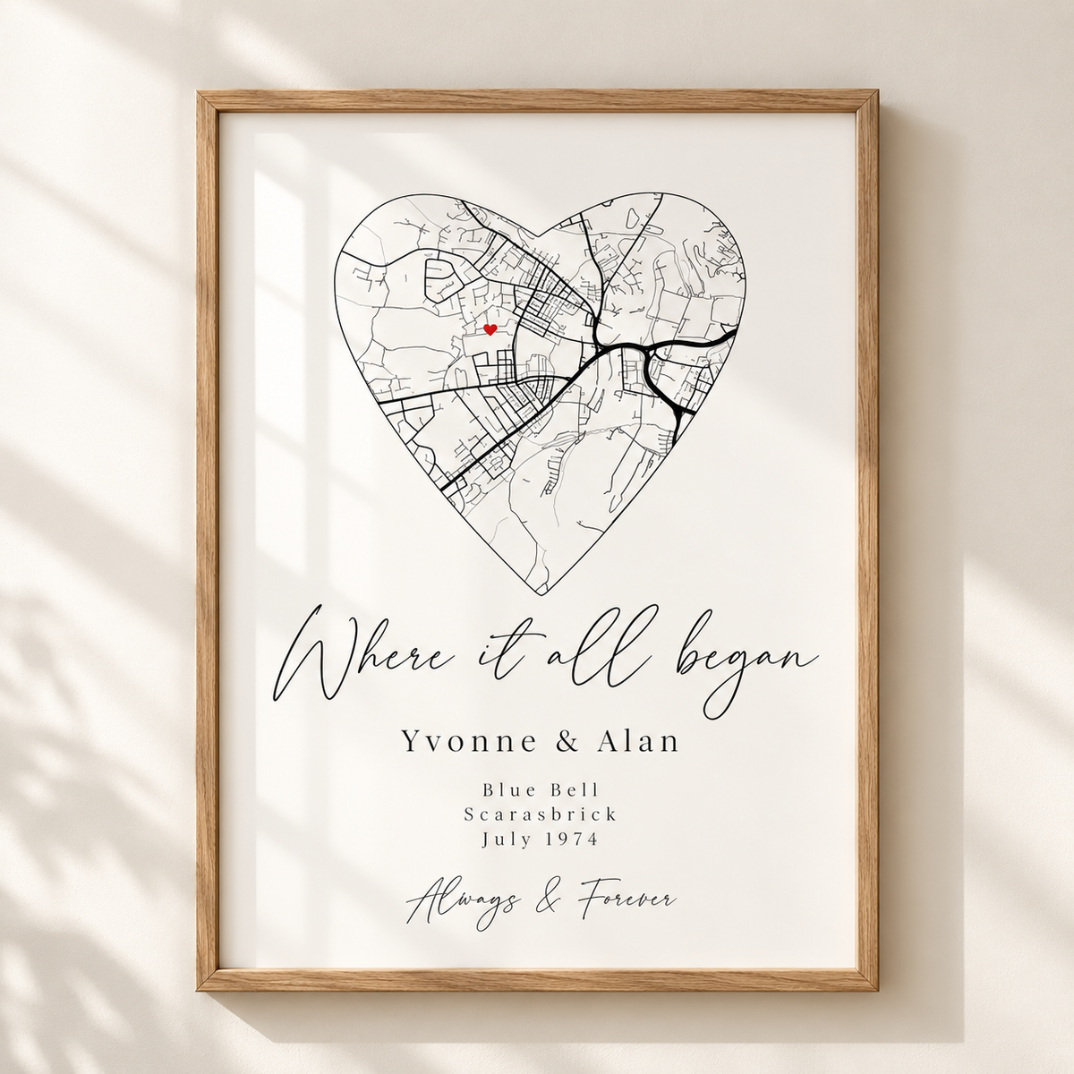 A personalized framed map print featuring a heart-shaped street map with a small red dot, the phrase 'Where it all began,' and the names 'Yvonne & Alan' with wedding details below, shown in a light wood frame on a white wall with soft shadow overlays.