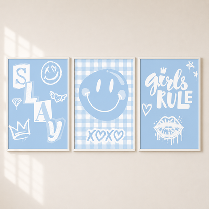 Three framed light blue and white decorative wall art prints hang side by side on a beige wall. The left print says "SLAY" with smiley faces and crown motifs, the center print shows a large smiley face on a gingham background with "XOXO" below, and the right print reads "girls RULE" with stars and a lip graphic.