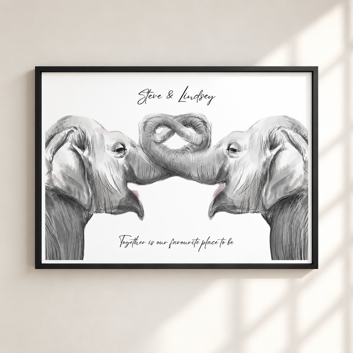 A framed personalized elephant art print hanging on a light beige wall. The print features a black and white sketch of two elephants facing each other with their trunks forming a heart shape, with the text 'Steve & Lindsay' at the top and 'Together is our favourite place to be' at the bottom.