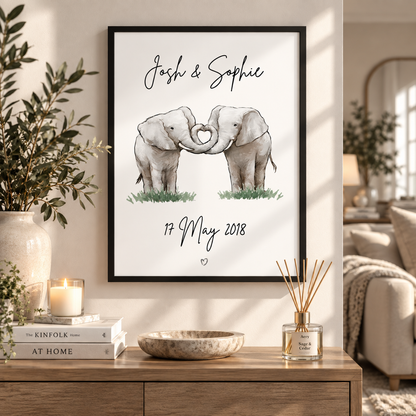 A framed personalized elephant art print hangs on a beige wall in a styled living room setting. The black-framed print features a watercolor illustration of two gray elephants forming a heart shape with their trunks, with the names 'Josh & Sophie' and the date '17 May 2018' written above and below.