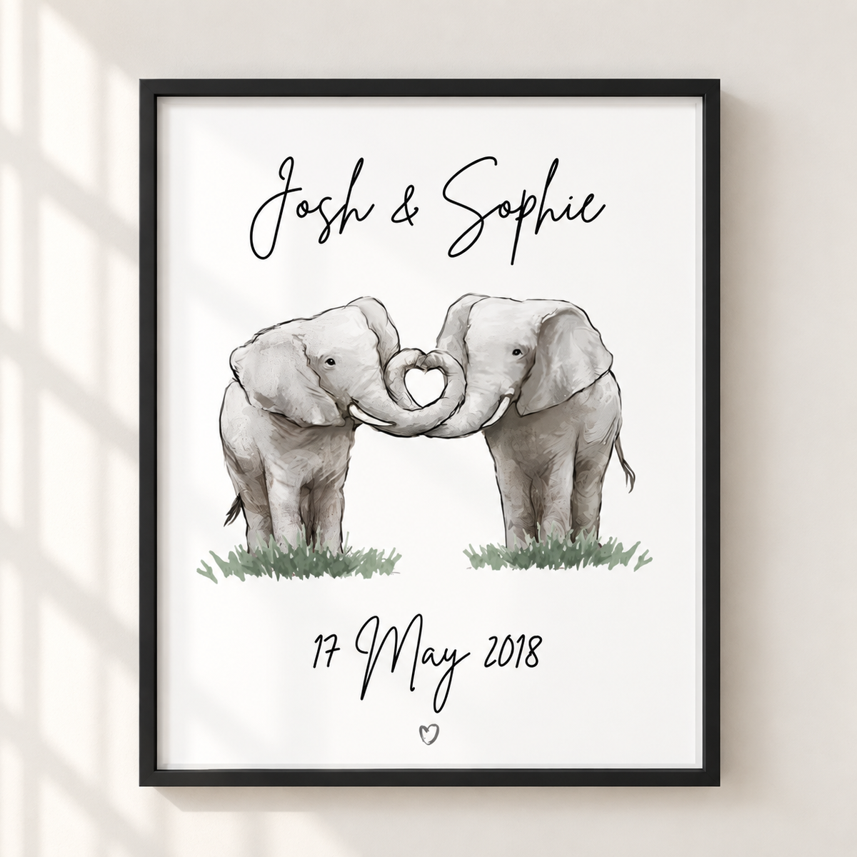 A framed personalized elephant art print hanging on a light beige wall. The print features two watercolor baby elephants forming a heart shape with their trunks, with the names 'Josh & Sophie' at the top and the date '17 May 2018' at the bottom.