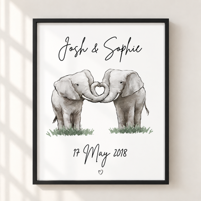 A framed personalized elephant art print hanging on a light beige wall. The print features two watercolor baby elephants forming a heart shape with their trunks, with the names 'Josh & Sophie' at the top and the date '17 May 2018' at the bottom.