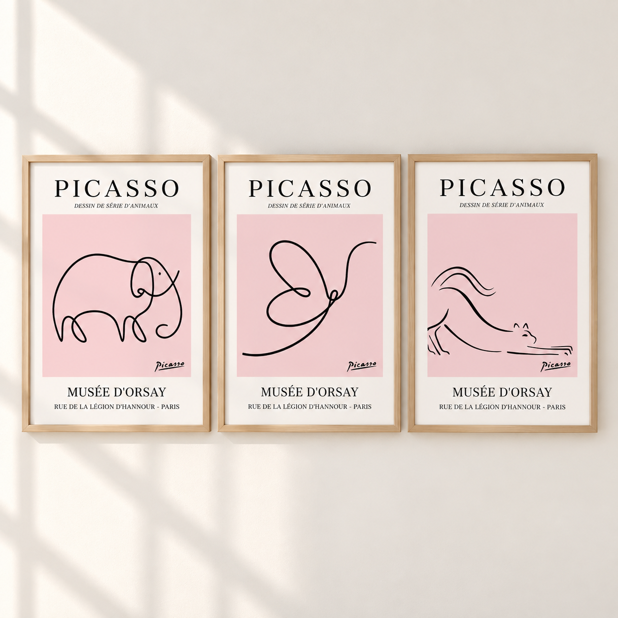 Three framed minimalist Picasso art prints hang side by side on a light wall with soft shadow overlays. Each print has a light pink square background, black line drawings (elephant, butterfly, horse), and white text reading PICASSO and MUSÉE D'ORSAY.