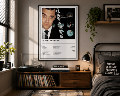 A framed Robbie Williams 'I've Been Expecting You' music poster hanging on a bedroom wall above a wooden record cabinet, with a bed, lamp, and plants in the room.