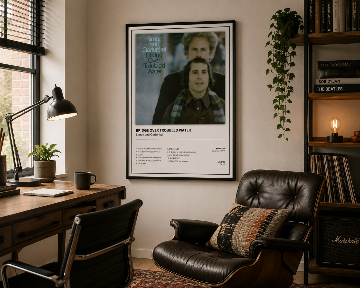 A framed music poster for Simon Garfield and Bridge of Broken Water hangs on a beige wall in a styled home office. The room features a wooden desk with a black desk lamp, a black leather armchair with a patterned pillow, a bookshelf with books and a hanging plant, and a window on the left.