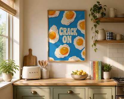 A framed wall art print featuring a blue background with illustrated fried eggs and the words “CRACK ON” in large cream letters, displayed in a bright kitchen above a countertop with a toaster, bowl of lemons, and plants.