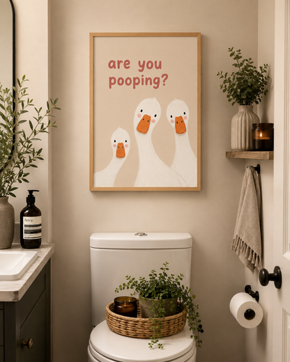 A framed wall art print featuring three cartoon white ducks with orange beaks and the text 'are you pooping?' in red, displayed above a toilet in a styled bathroom setting.