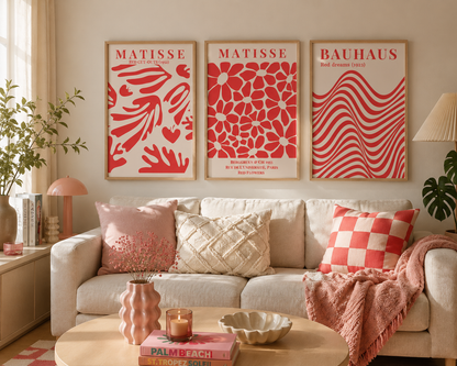 Three framed Matisse and Bauhaus art prints in red and cream hang above a beige sofa in a styled living room with pink pillows, a pink throw blanket, and a round coffee table.