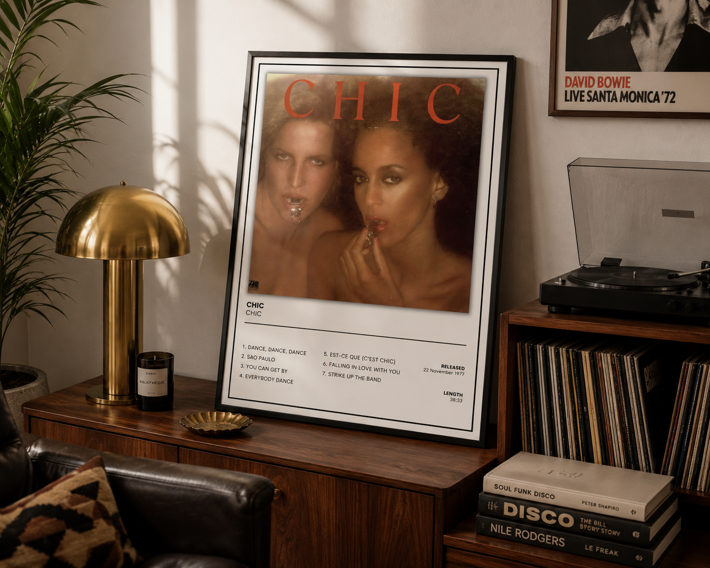 A framed CHIC magazine cover print leaning on a wooden sideboard in a styled room with a brass lamp, records, and a record player.