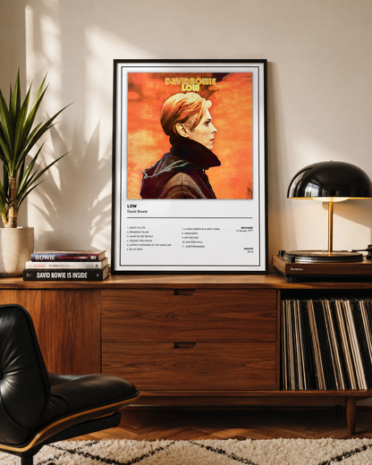 A framed music poster for David Bowie's 'Low' is displayed on a wooden credenza in a modern living room setting, flanked by a potted plant, a black table lamp, and a black leather chair.