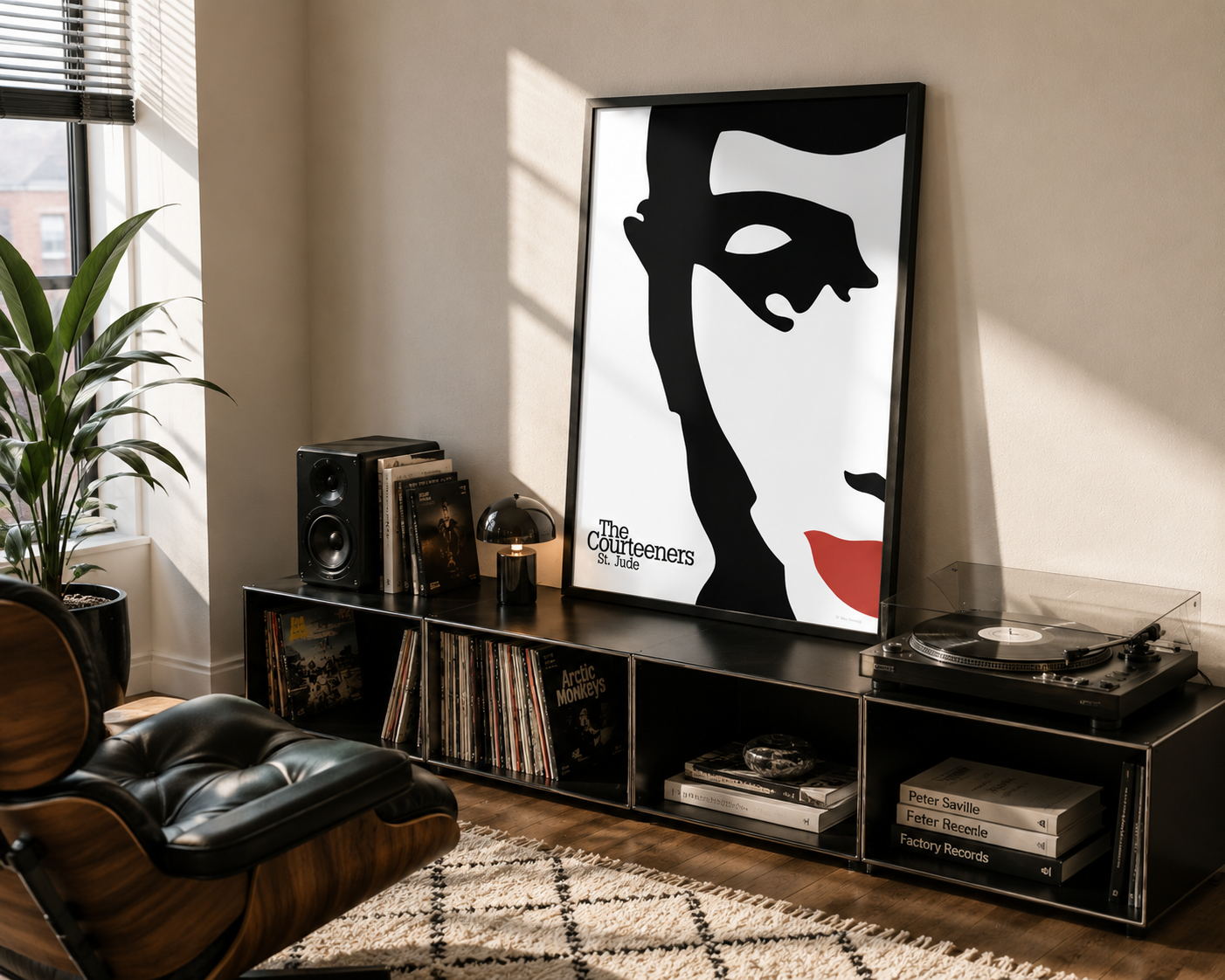 A framed black-and-white poster with a bold red lip accent and the text 'The Courteeners' leaning on a black media console in a styled living room. The room includes a leather lounge chair, speakers, records, a turntable, and a plant.