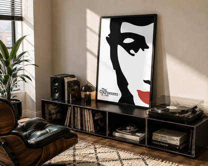A framed black-and-white poster with a bold red lip accent and the text 'The Courteeners' leaning on a black media console in a styled living room. The room includes a leather lounge chair, speakers, records, a turntable, and a plant.