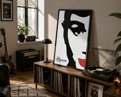 A large framed poster of The Courteeners 'St Jude' leaning on a wooden record cabinet in a styled room with a record player, lamp, guitar, and plants.