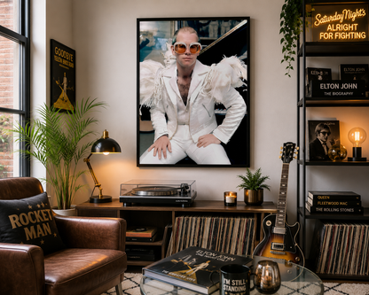 A large framed wall art print depicting Elton John in a white feathered suit and sunglasses hangs on a wall in a styled living room with a leather chair, plants, a turntable, and an Elton John themed shelf.