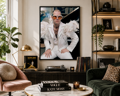 A large framed vertical art print of Elton John in a white suit hangs on a beige wall in a styled living room with a pink armchair, green sofa, and coffee table.