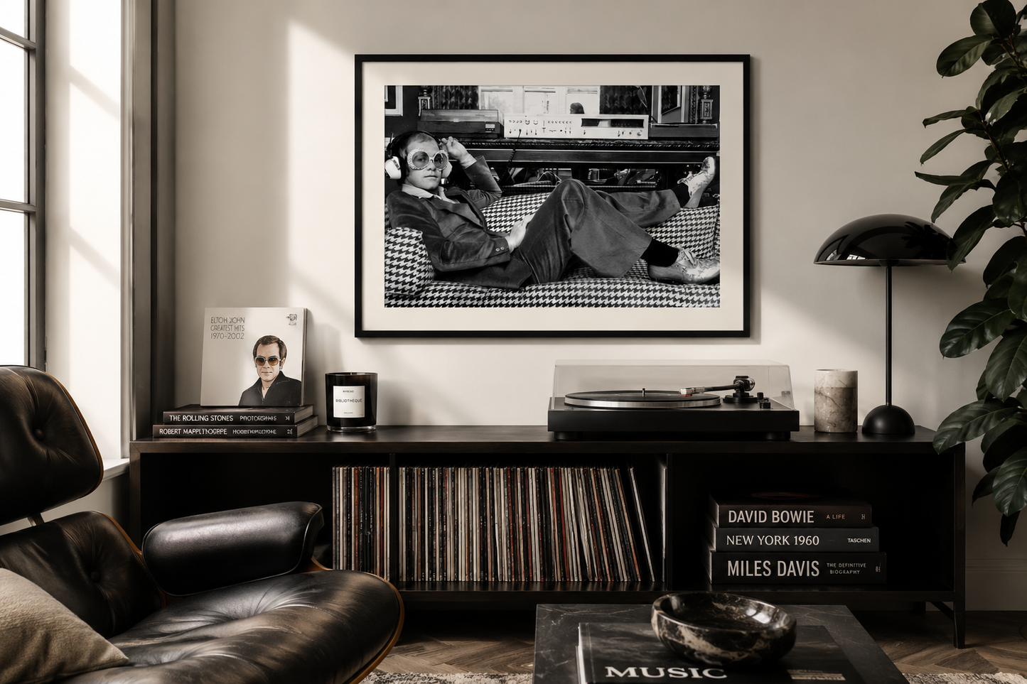 A framed black-and-white art print of Elton John lounging on a couch, displayed on a wall above a dark media console in a modern living room setting.