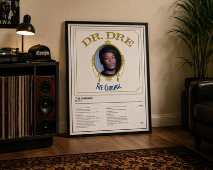 A framed music poster for Dr. Dre's 'The Chronic' leaning against a beige wall in a styled room with a record rack, lamp, plant, and chair.