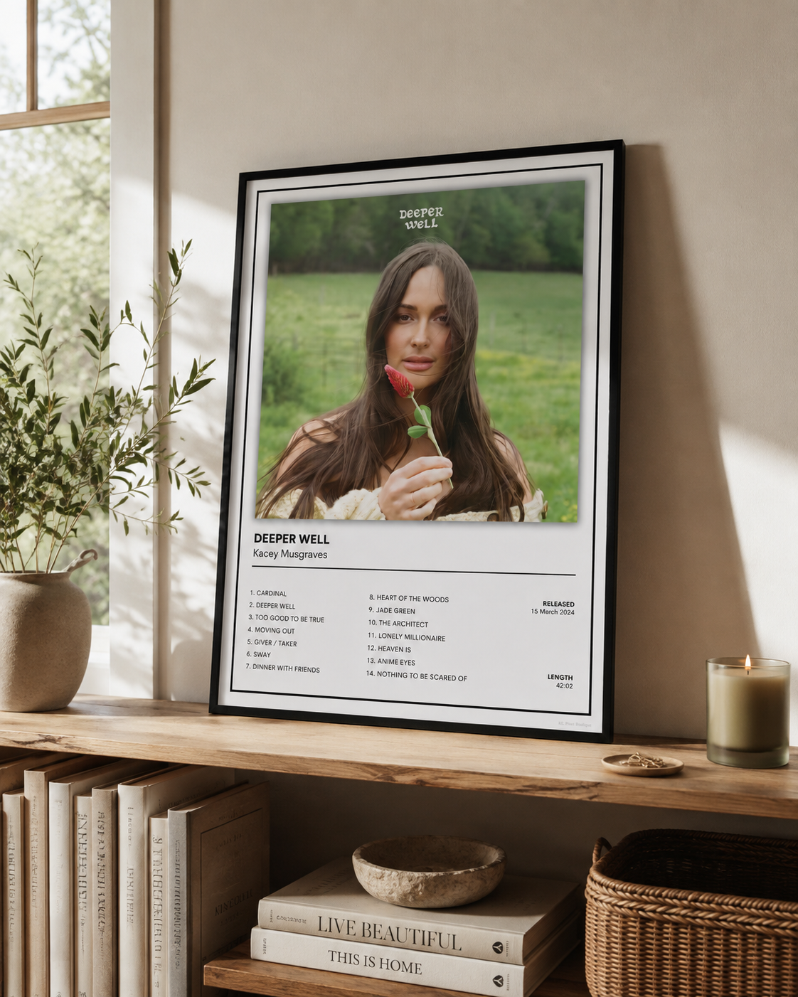 A framed art print featuring a portrait of Deeper Well with song lyrics below, leaning on a wooden shelf in a styled home interior next to a window, books, and a candle.