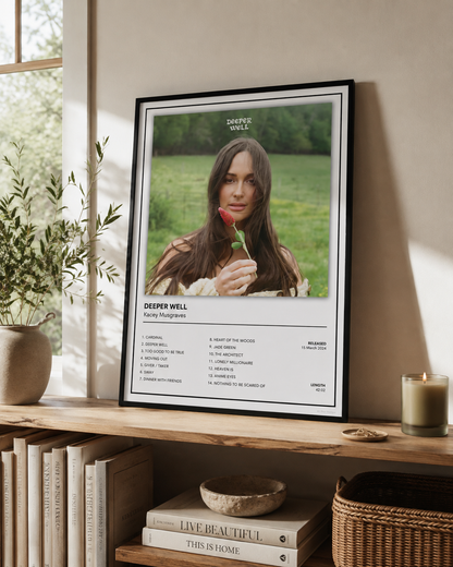 A framed art print featuring a portrait of Deeper Well with song lyrics below, leaning on a wooden shelf in a styled home interior next to a window, books, and a candle.