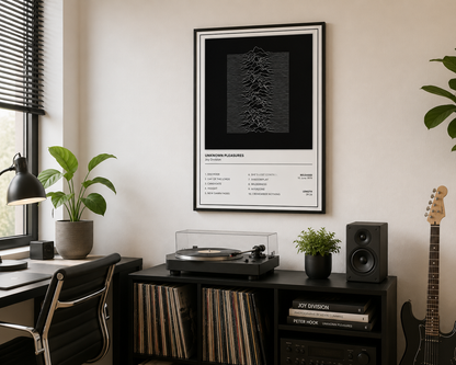 A framed black and white art print of Joy Division's 'Unknown Pleasures' hanging on a beige wall in a modern, styled music room with a turntable, speakers, and plants.