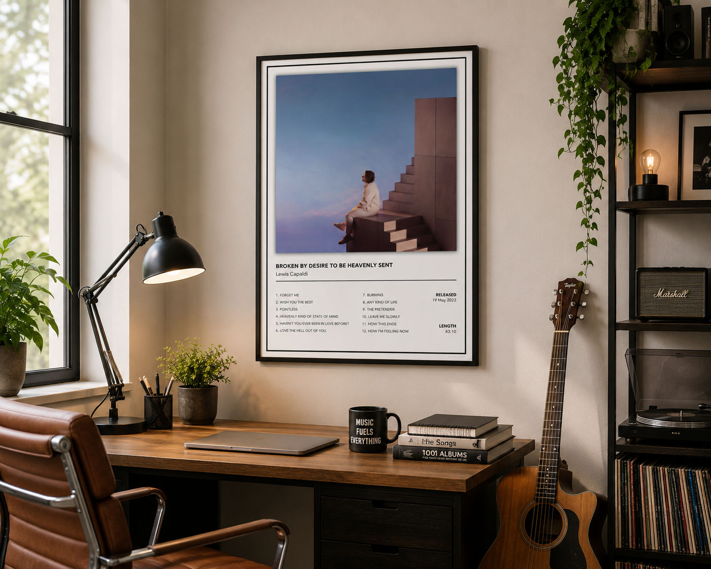 A framed art print of Lewis Capaldi poster, Broken By Desire To Be Heavenly Sent poster and text below, displayed on a wall above a wooden desk in a styled home office setting with a guitar, lamp, and plants.