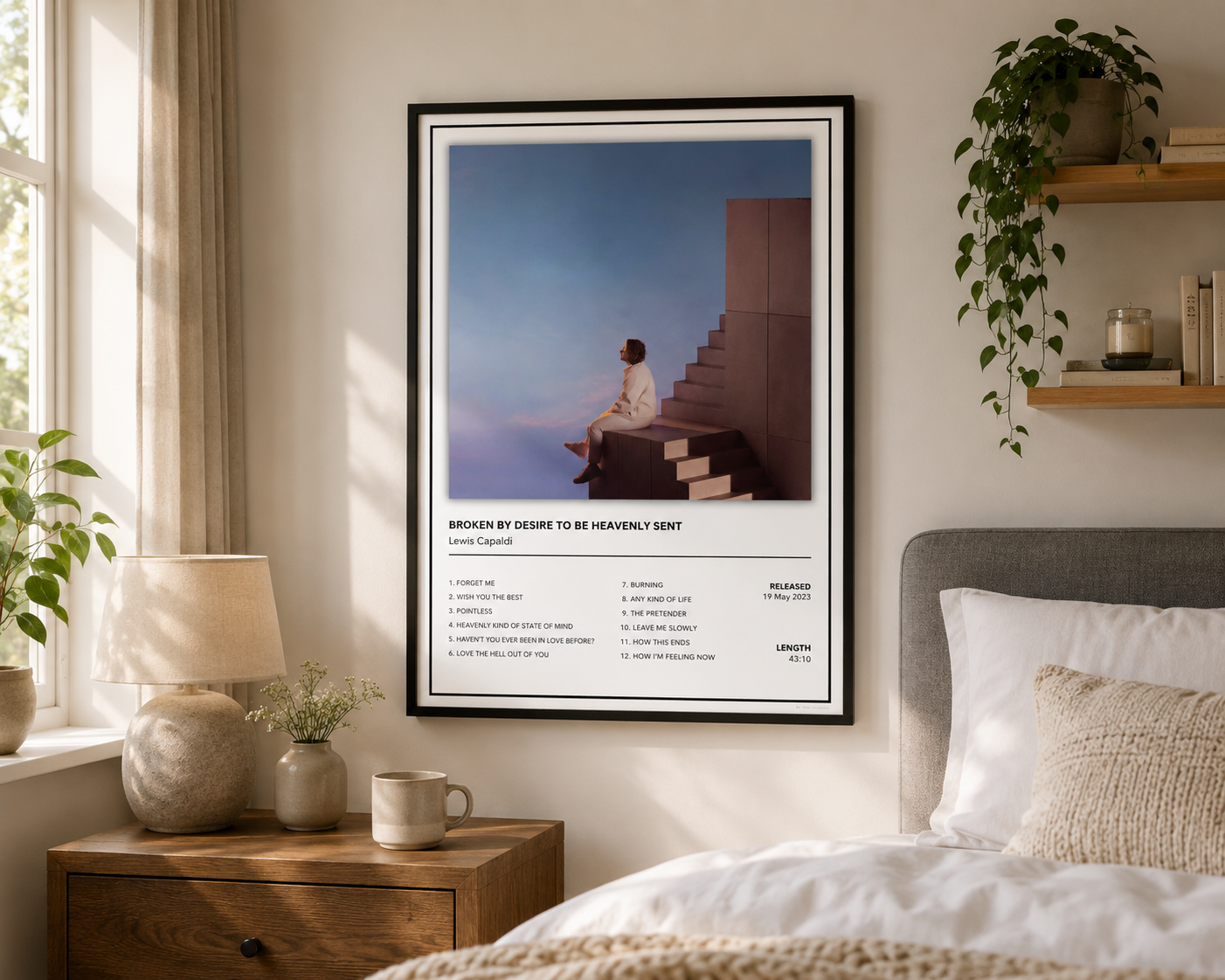 A framed art print featuring Lewis Capaldi, Broken By Desire To Be Heavenly Sent poster, displayed on a bedroom wall above a bed with plants and a bedside table.