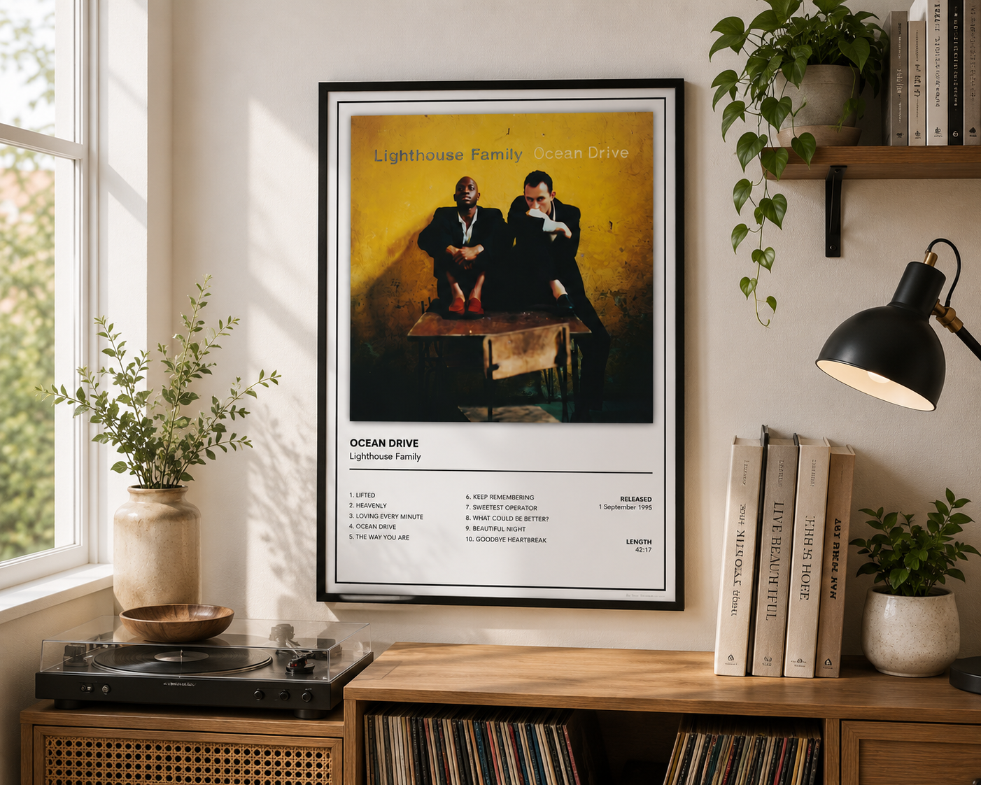 A framed Ocean Drive Lighthouse Family album poster hangs on a beige wall in a styled room setting with a wooden shelf, plants, books, and a record player.