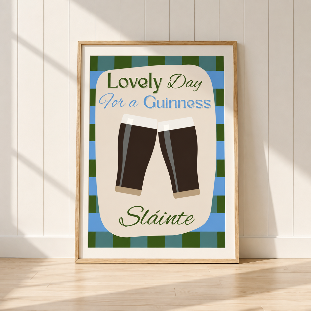 A framed art print leaning against a white paneled wall on a light wood floor. The print features a green and blue tartan border, a beige center, two clinking dark stout pint glasses, and the text 'Lovely Day For a Guinness Slainte'.