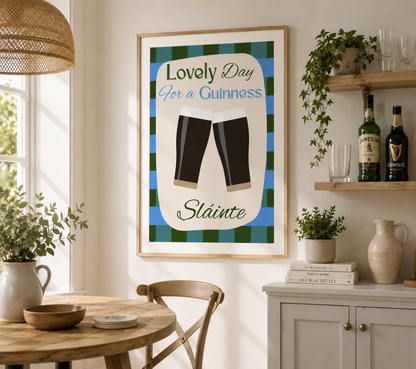 A framed wall art print hanging in a bright kitchen or dining area. The print reads “Lovely Day for a Guinness” and “Stainte” with two Guinness pint glasses centered on a green and blue plaid background.