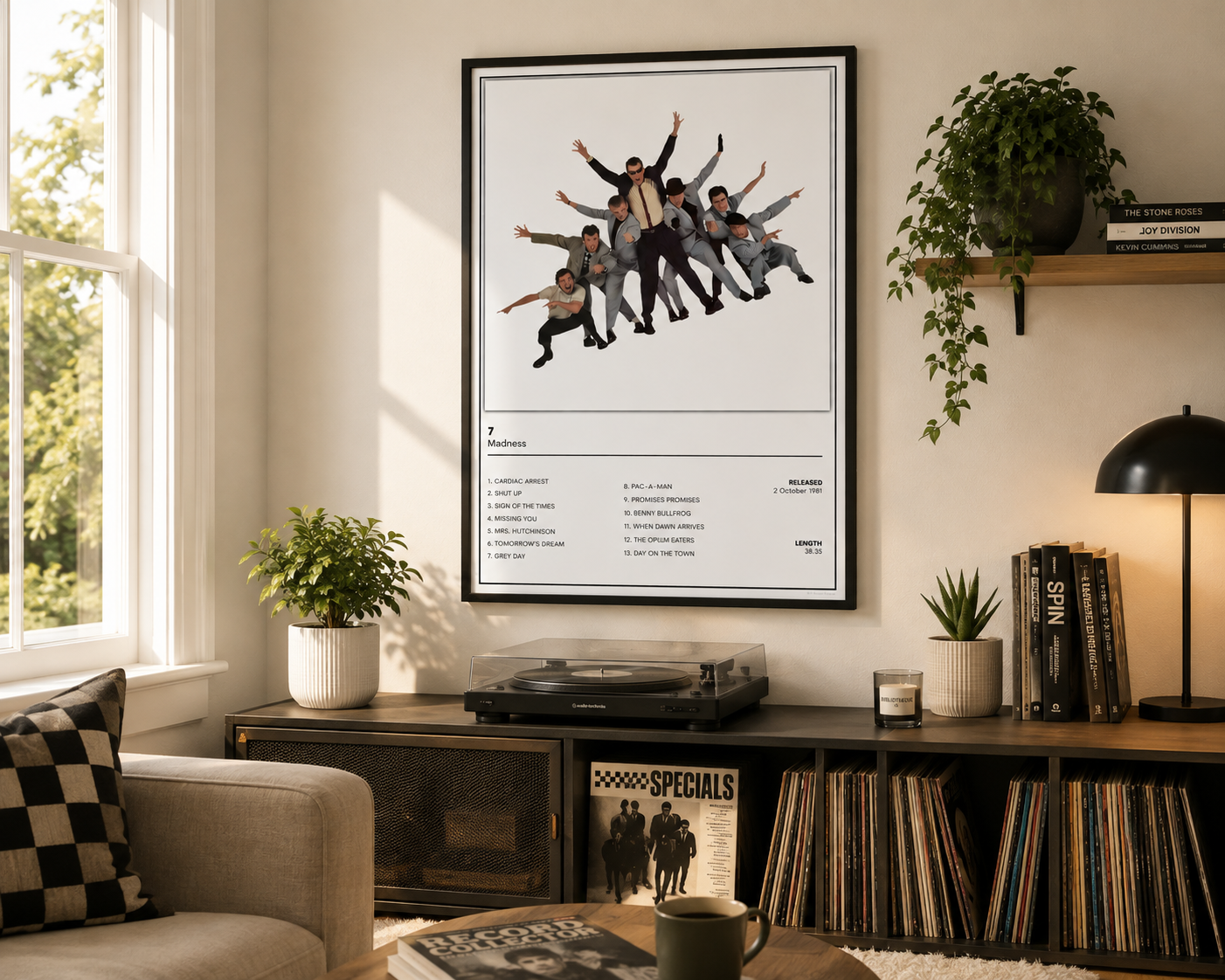A framed music poster of the band Madness 7 Album Poster hangs on a wall in a styled living room setting with a record player, books, plants, and a lamp.