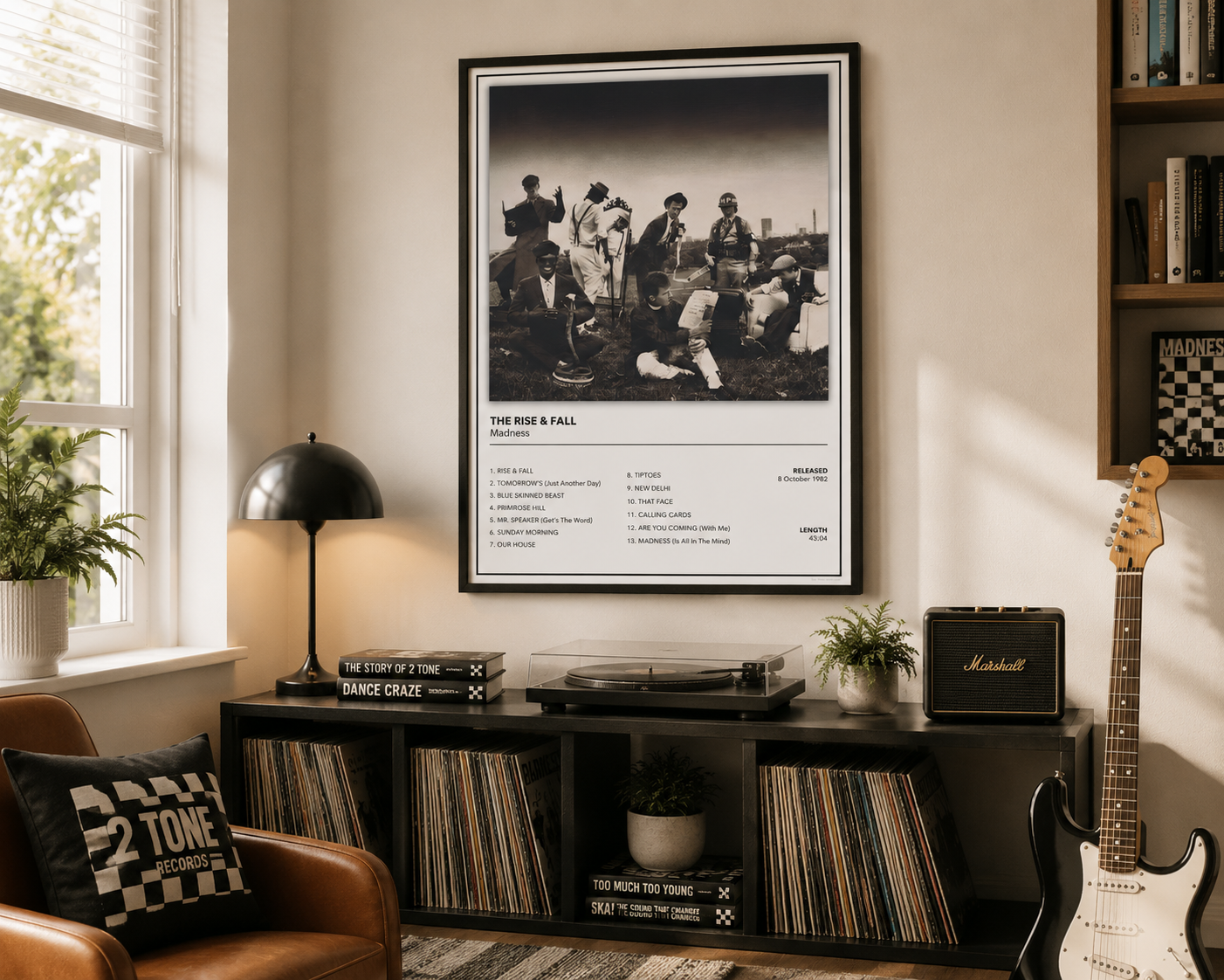A framed black-and-white music poster of Madness, hangs on a beige wall in a styled room with a record console, guitar, lamp, and shelf.