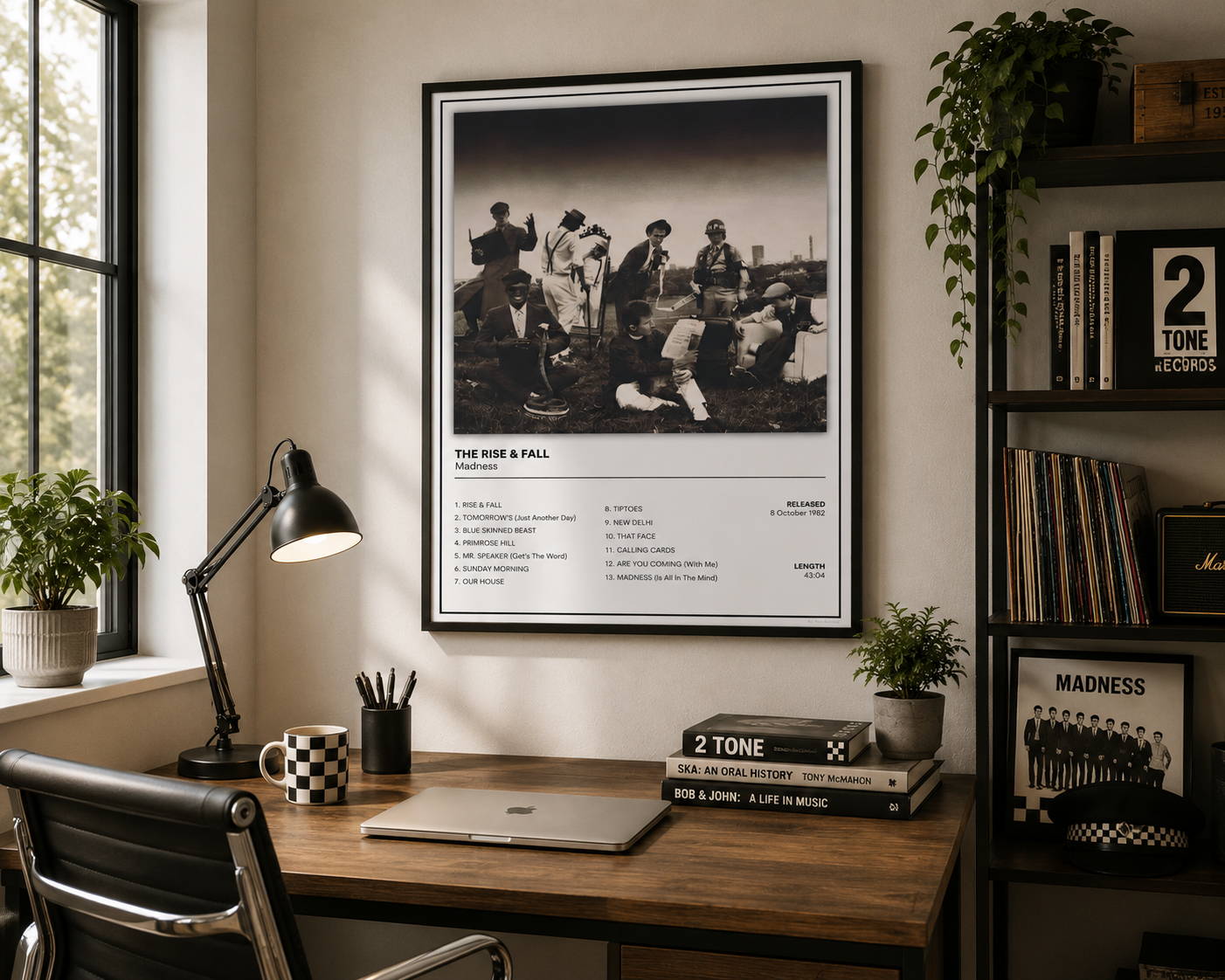 A framed black-and-white music poster for 'The Rise & Fall' hangs on a wall above a wooden desk in a styled home office setting with a lamp, laptop, plants, and record shelves.