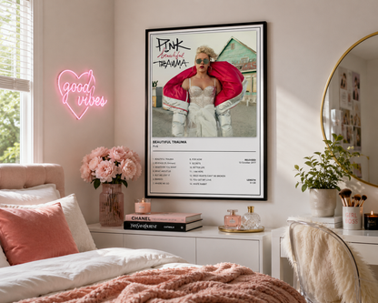 A framed 'Pink's album 'Beautiful Trauma' art print hangs on a bedroom wall above a bed, styled with a pink neon 'good vibes' sign, flowers, candles, and a mirror.