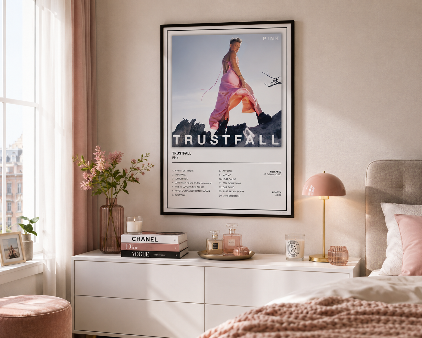 A framed fashion poster for Trustfall Pink displayed on a bedroom wall above a white dresser, styled with a pink lamp, books, and flowers.
