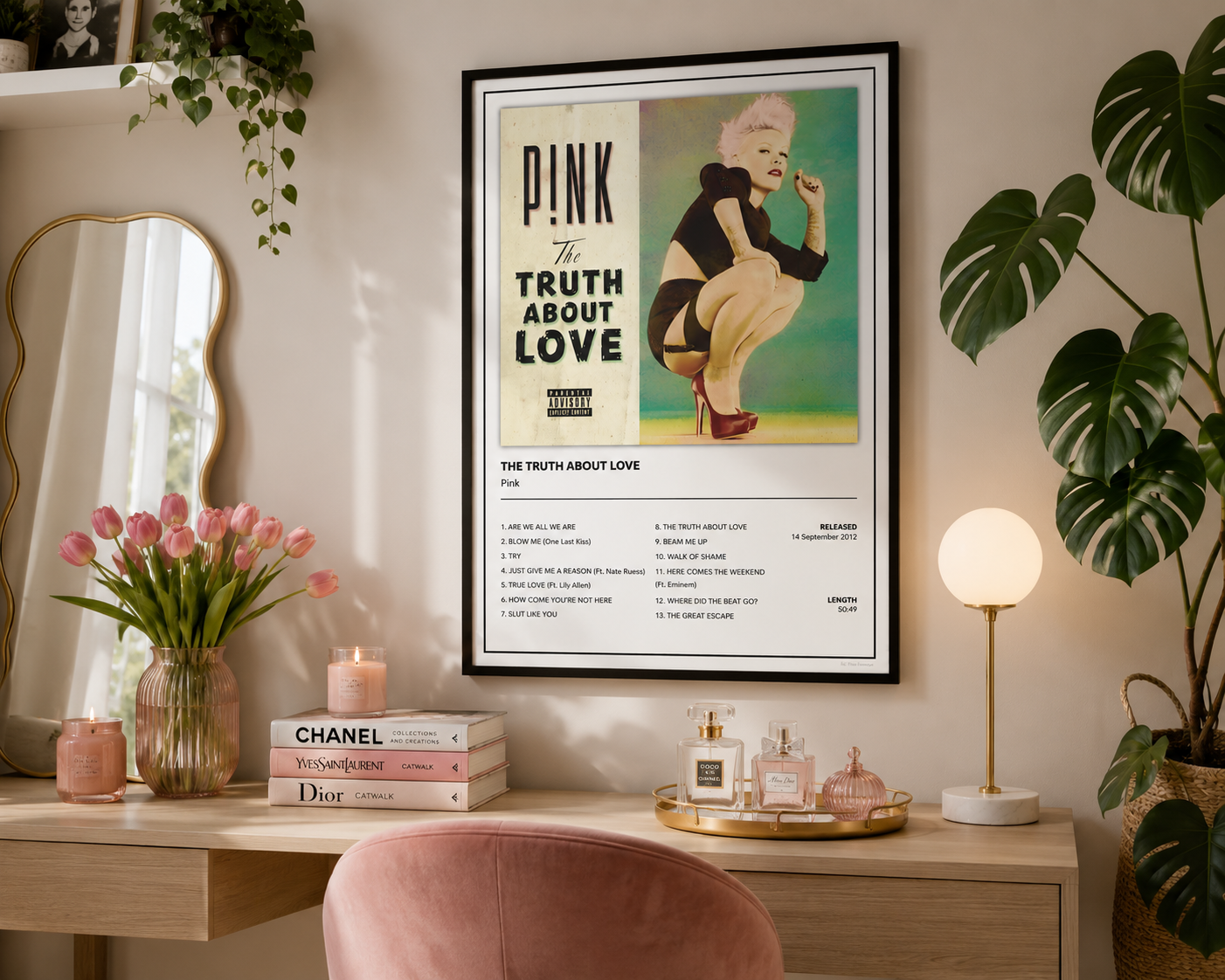 A framed P!NK 'The Truth About Love' poster hanging on a beige wall above a wooden vanity table in a styled room. The scene includes pink tulips, a mirror, a lamp, and potted plants.