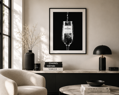 A framed black and white art print of a Chanel champagne flute hanging on a beige wall in a modern, sunlit living room setting.