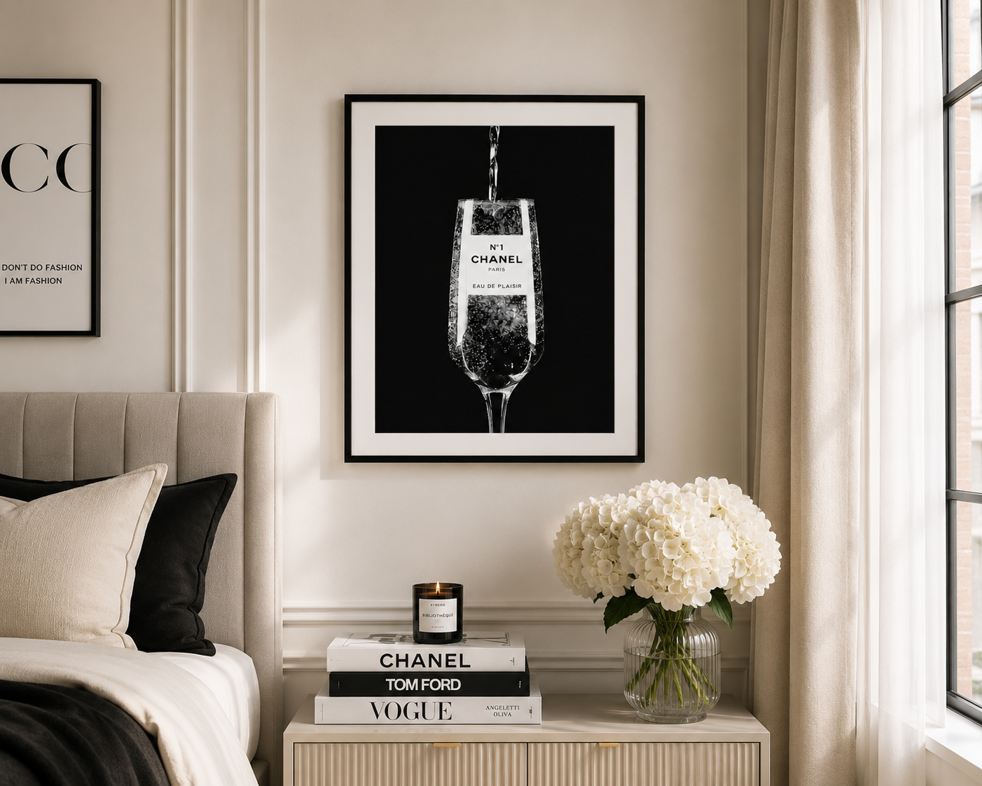 A framed black and white art print of a champagne flute with a Chanel logo, displayed on a beige wall in a styled bedroom setting above a nightstand.