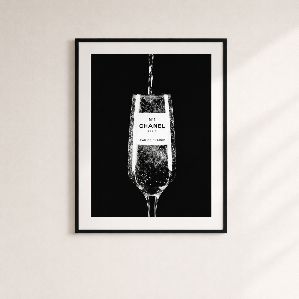 A framed black and white art print featuring a champagne flute with the text 'No.1 CHANEL PARIS' and 'EAU DE FLORIS' on the label, hanging on a light beige wall.