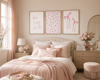 A styled bedroom interior with a bed dressed in blush pink and cream bedding, three framed wall art prints above the headboard, matching pink flowers on bedside tables, and a soft blush-toned room overall.
