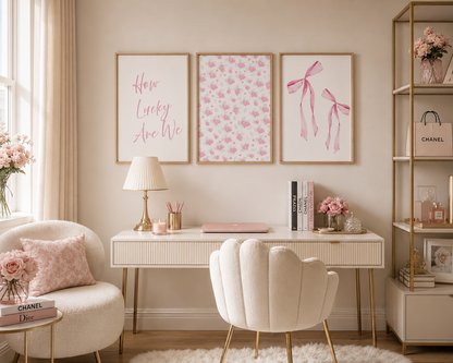A styled home office interior with three framed wall art prints above a white desk and cream shell chair. The prints feature pink script text, pink floral patterning, and pink ribbon illustrations.