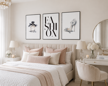 A styled bedroom interior with a bed dressed in cream and blush bedding, three framed wall art prints above the headboard, matching pillows, bedside tables with lamps, a round mirror, and a blush velvet chair.