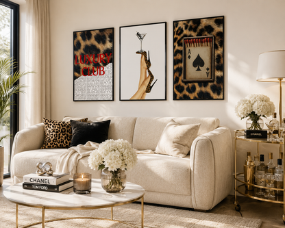 Three framed wall art prints with leopard print and fashion-themed designs hang above a cream sofa in a styled living room. The prints feature a red 'LUXURY CLUB' text on the left, a high-heel graphic in the center, and an aces de pied card on the right.
